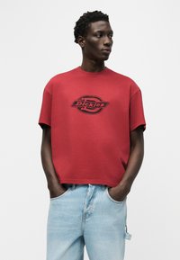 Άνδρας που φοράει κόκκινο t-shirt με το λογότυπο Dickies και ανοιχτόχρωμο τζιν, με τα χέρια στις τσέπες, κοιτάζει προς τα αριστερά του μπροστά σε απλό φόντο.