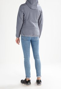 Veste à capuche grise avec une texture lisse, un design ajusté et des détails de couture visibles, associée à un jean bleu clair et des baskets noires.