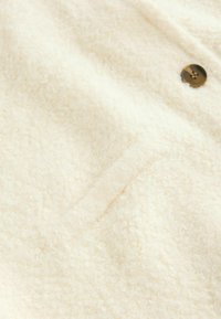 Tissu bouclé de couleur crème douce avec une petite poche fendue et un seul bouton visible en marron foncé.