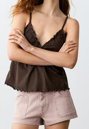 Femme aux bras croisés portant un haut sans manches marron foncé et un short rose clair clouté, sur un fond uni.