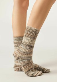 Chaussettes à carreaux marron et beige avec des bords côtelés, présentant une texture douce et des rayures variées, affichées sur des pieds allant de la cheville au mollet.