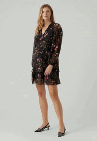 Vestido preto com estampado floral, tecido transparente, decote em V, mangas longas, bainha em camadas e padrões de flores cor-de-rosa. Combinado com sapatos de salto agulha pretos.