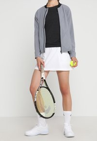 Grå atletisk jacka, svart tröja, vit kjol och vita tennisskor. Håller en tennisboll och racket. Enkla mönster och lätta material.