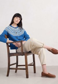 Sininen neulejakku, jossa on valkoinen Fair Isle -kuvio, puunappilukko, kermaovet ja ruskeat liukukengät, istuu puisella tuolilla.