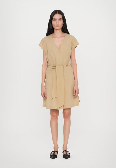 Robe portefeuille beige avec des manches courtes, un décolleté en V et une ceinture à nouer ; elle présente une jupe fluide et des poches latérales. Portée avec des chaussures plates noires.