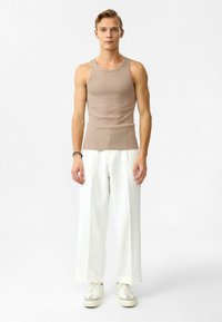 Beige geribbelde tanktop gecombineerd met witte wijde broek en witte sneakers. Model staat rechtop tegen een effen achtergrond.