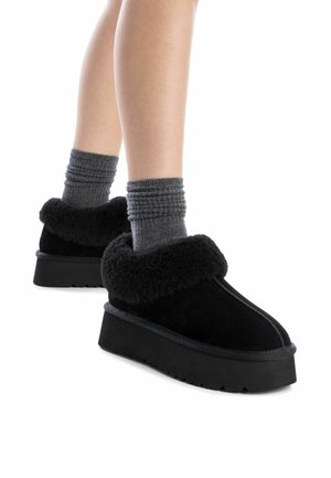 Zapatos de ante negros tipo slip-on con una plataforma negra gruesa y texturizada y puños de peluche negros, combinados con calcetines grises de canalé.