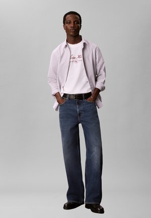 Junger Mann steht mit den Händen in den Taschen, trägt ein gestreiftes Hemd über einem weißen T-Shirt mit Logo, weite blaue Jeans, schwarzen Gürtel und schwarze Schuhe.