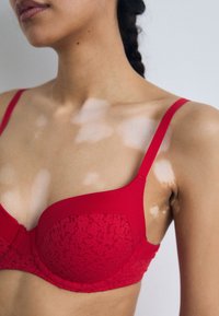 Crvena bralette s čipkastom teksturom, glatkim materijalom, podesivim naramenicama i oblikom koji se prilagođava. Ima suptilan sjaj i dekorativno šivanje.