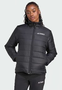 Γυναίκα ντυμένη με μαύρη τζάκετ Terrex με πλισέ και ταιριαστά παντελόνια, στέκεται με τα χέρια στις τσέπες ενάντια σε φωτεινό φόντο.