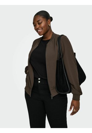 Smilende kvinde med håret sat op i en knold, iført en brun lynlåstrøje, sort top, sorte bukser, gyldne creoløreringe, ringe og bærende på en stor sort taske.