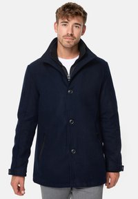 Manteau en mélange de laine bleu marine avec un col haut, une fermeture à boutons et deux poches latérales zippées. Présente une texture lisse et un design ajusté.