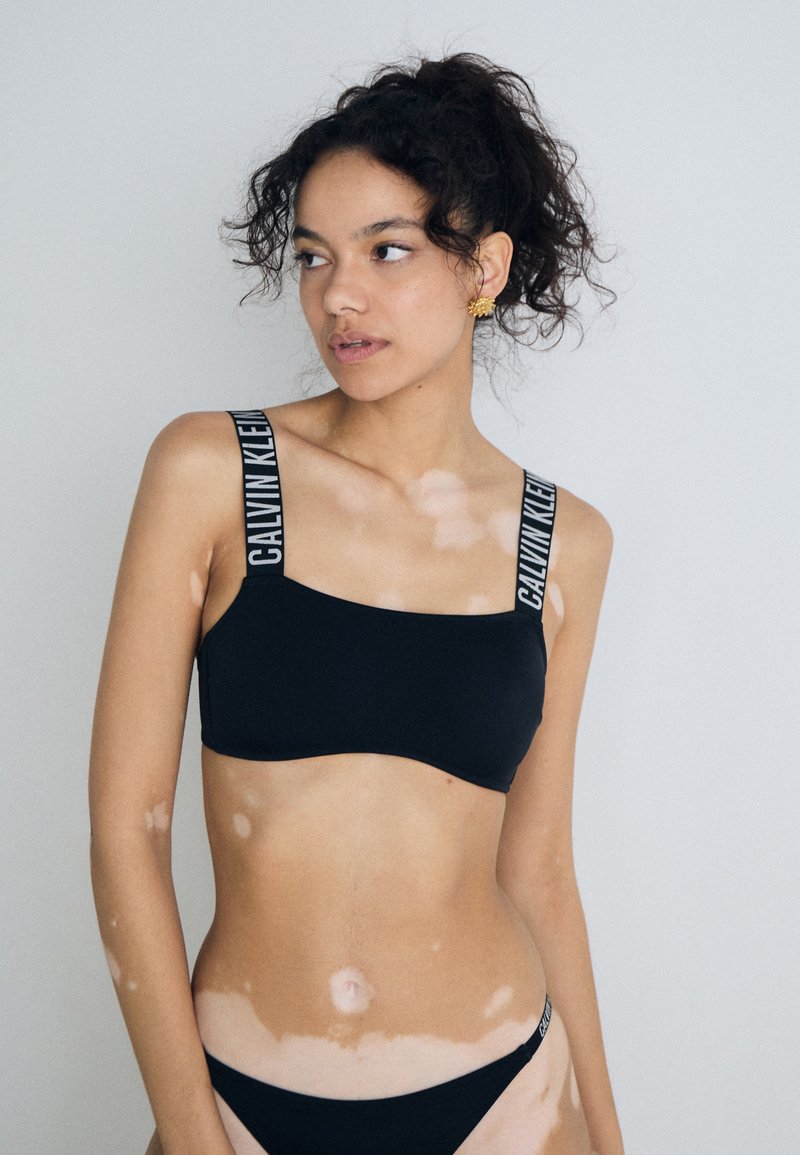 Schwarzer Bikini-Oberteil mit breiten Trägern, das mit dem "CALVIN KLEIN"-Logo versehen ist. Glatte Textur, minimalistisches Design, quadratischer Ausschnitt und passende Unterteile.