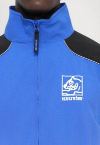 Gros plan sur une personne portant une veste à fermeture éclair bleue avec des panneaux noirs aux épaules et un logo blanc "Icecream" représentant un visage animal stylisé.