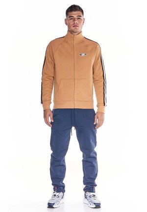 Un homme pose avec une veste de survêtement beige zippée, ornée de bandes sombres sur les manches, un pantalon de jogging bleu et des baskets blanches, sur fond blanc.