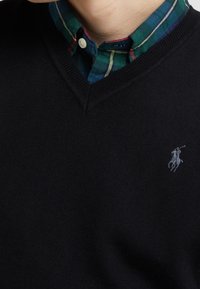 Black knitted sweater with a V-neck design, featuring a blue and green plaid collared shirt underneath and a small gray logo on the chest.