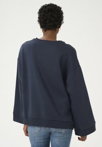 Navy zilgans izmēra džemperis ar nolaistām plecu šuvēm un ribotām piedurknēm, kombinēts ar gaiši ziliem džinsiem. Vienkārša dizaina, mīksta tekstūra.