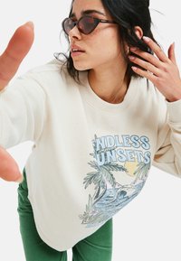 Valkoinen teksturoitu collegepaita, jossa on graafinen printti palmuista ja auringosta, yhdistettynä vihreisiin corduroyhousuihin ja tummiin sydämenmuotoisiin aurinkolaseihin.