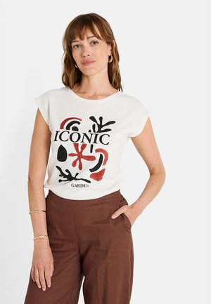 Camiseta blanca de manga corta con un estampado gráfico en negro y rojo, que presenta la frase "JARDÍN ICÓNICO". Combinada con pantalones anchos marrones.