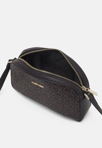 Braune und schwarze Calvin Klein Handgelenktasche mit Monogrammmuster, goldener Reißverschluss und Logo, teilweise geöffnet auf weißem Hintergrund.