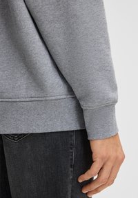 Primo piano di una mano di una persona appoggiata accanto all'anca, che indossa un felpa grigia e jeans di denim neri.