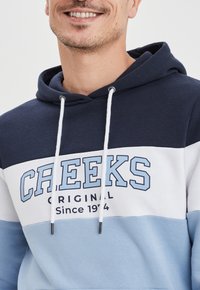 Sudadera con capucha en un diseño de bloques de color azul marino, blanco y azul claro. Presenta la palabra "CREEKS" impresa en azul claro y una capucha con cordón.