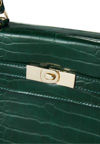 Détail d'un sac à main vert en cuir synthétique façon crocodile avec fermoir rotatif rectangulaire doré et bords cousus.