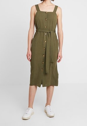 Vestito verde oliva, di lunghezza al ginocchio, con scollatura quadrata, spalline larghe, bottoni frontali, tasche laterali e una cintura abbinata. Abbinato a sneakers bianche.