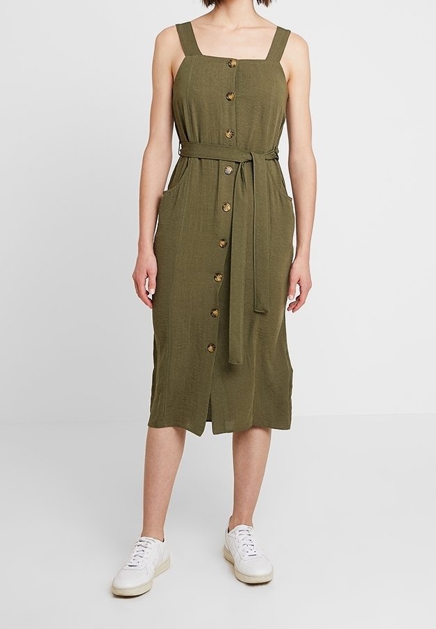 Vestito verde oliva, di lunghezza al ginocchio, con scollatura quadrata, spalline larghe, bottoni frontali, tasche laterali e una cintura abbinata. Abbinato a sneakers bianche.