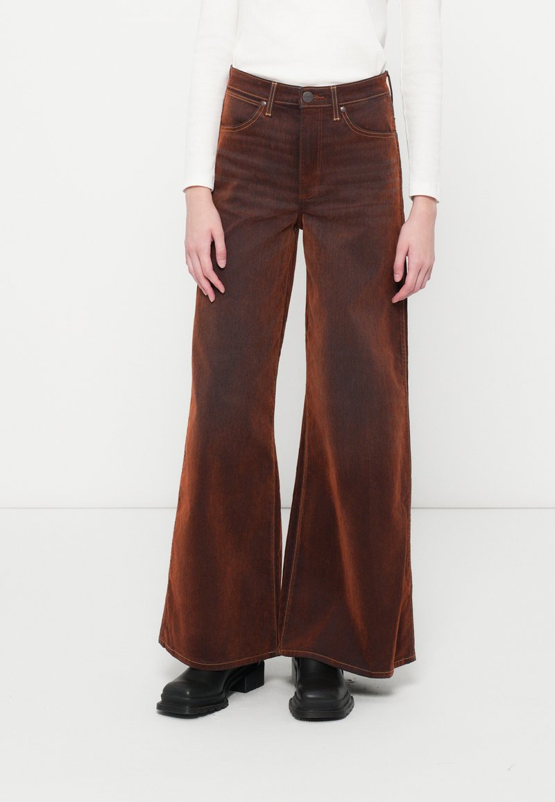 Persona che indossa jeans marroni a vita alta e larghi, insieme a stivali neri chunky, con una maglietta bianca a maniche lunghe, su uno sfondo bianco semplice.