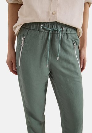 Osoba nosząca oliwkowe spodnie ze sznurkiem i srebrnymi kieszeniami na zamek oraz beżową koszulę zapiętą i wpuszczoną z przodu.