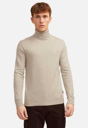 Jeune homme aux cheveux courts et bouclés portant un pull à col roulé beige et un pantalon foncé, debout devant un fond clair uni.