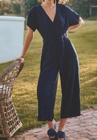 Marineblå, stribet jumpsuit med korte ærmer, omslagdesign og en bindebælte i taljen. Kombineret med blå slippers med blomsterdetaljer.