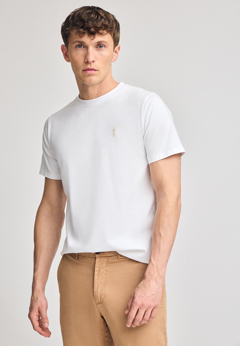 Vit bomull t-shirt med rund halsringning, korta ärmar och en liten beige broderad logotyp på vänster bröst. Matchad med beigefärgade byxor.