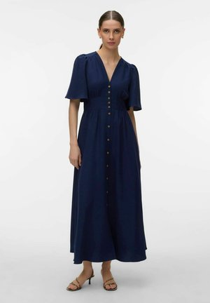 Nő sötétkék, gombos, hosszú ruhában, rövid, fodros ujjakkal és nyitott orrú magassarkú szandálban, egyszínű fehér háttér előtt.