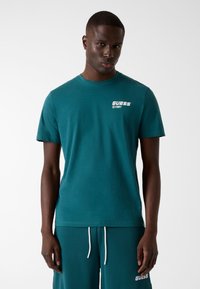 Teal cotton t-shirt with a crew neckline, featuring a white "GUESS" logo and 1981 detail on the left chest. Simple, short-sleeved design.