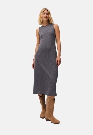 Mujer de pie con un vestido midi gris sin mangas de punto acanalado y botas vaqueras marrones de ante sobre un fondo liso.