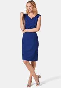 Blaues ärmelloses Kleid mit V-Ausschnitt, tailliertem Design und knielanger Länge. Kombiniert mit hellgrauen Riemchen-Absätzen.