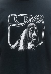 Must-valge graafikuga must särk, millel istuva basseetkoera kujutis ja stiliseeritud tekst, mis moodustab koera kohal ristküliku kujundi.