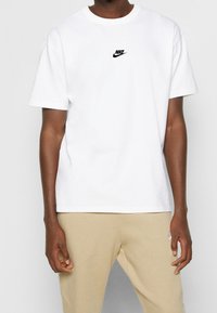 Bílý bavlněný tričko s malým černým logem Nike na hrudi. Kulatý výstřih, krátké rukávy, volný střih.