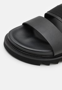 Sandale noire à glissière avec une finition mate, dotée de deux larges sangles et d'une semelle texturée pour une meilleure adhérence. Design simple et minimaliste.