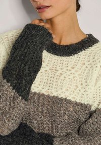 Frau in einem grob gestrickten Pullover mit dunkelgrauen, cremefarbenen und braunen Farbblöcken, die mit der Hand den Hals in der Nähe des Kragens berührt.
