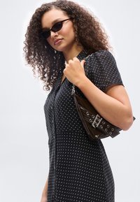 Γυναίκα με σγουρά μαλλιά, φορώντας μαύρα γυαλιά ηλίου και μαύρο φόρεμα με πουά, κρατώντας μια σκούρα καφέ τσάντα χειρός με μεταλλικές αγκράφες.