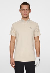 Beige polotröja med krage, korta ärmar och en svart logotyp på bröstet. Tillverkad av smidigt tyg, i kombination med mörka byxor och en vit handske.