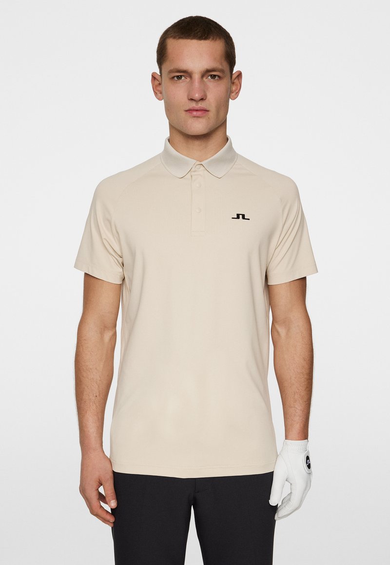 Beige polotröja med krage, korta ärmar och en svart logotyp på bröstet. Tillverkad av smidigt tyg, i kombination med mörka byxor och en vit handske.