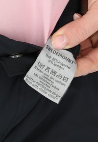 Schwarzes Kleidungsstück mit einem Pflegeetikett, das die Stoffzusammensetzung zeigt: 80 % Polyester, 12 % Spandex. Das Etikett enthält Waschsymbole und den Markennamen.