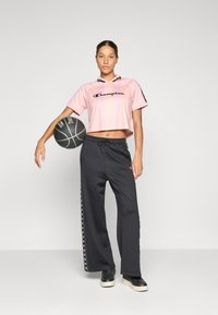 Jovem mulher vestindo uma camiseta cropped rosa da Champion e calças de treino pretas, posando com uma bola de basquete preta e branca contra um fundo simples.