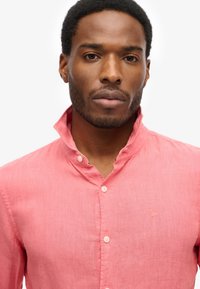 Lichtroze linnen shirt met een staande kraag en knoopsluiting. Bevat minimalistisch logo borduursel op de borst en subtiele textuur.