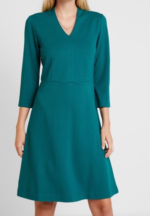 Woman wearing a teal knee-length dress with three-quarter sleeves and a V-neckline, standing against a plain light background.