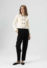 Veste courte couleur crème avec col rond et six boutons noirs, associée à un pantalon noir taille haute et des ballerines noires.
